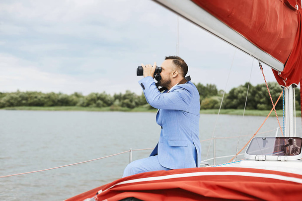 Young and brutal businessman with the beard in a stylish blue suit sits on the yacht ship and watching through the binoculars - Foto, Imagem