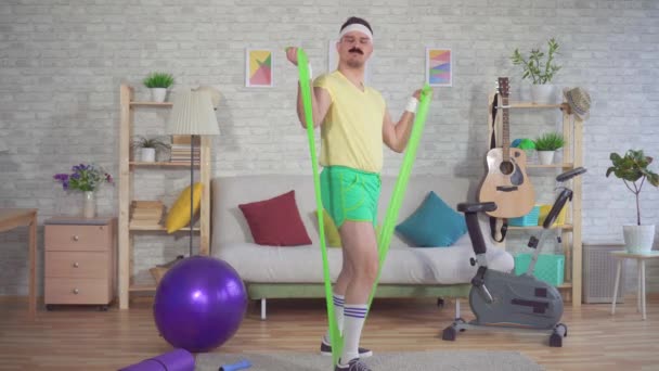 Funny athlete of the 80s with a mustache engaged in fitness at home with the help of an elastic band - Filmmaterial, Video