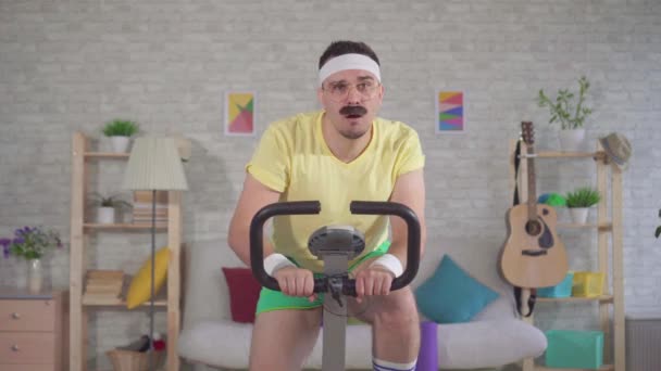 Portrait funny energetic man athlete from the 80s with a mustache engaged at home on a stationary bike - Filmmaterial, Video