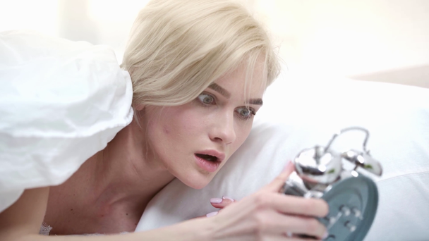 surprised blonde girl lying in bed, taking alarm clock and standing up  - Metraje, vídeo