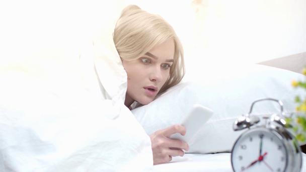 επιλεκτική εστίαση του συναγερμού κοντά ξανθιά έκπληκτη γυναίκα ξύπνημα και κοιτάζοντας smartphone στο υπνοδωμάτιο  - Πλάνα, βίντεο