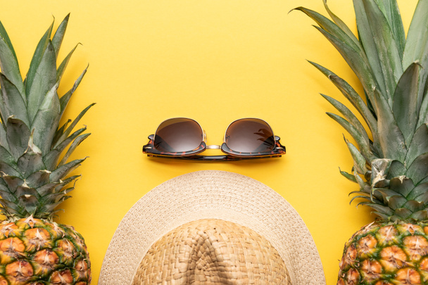 Top zicht op stro hoed en zonnebril bij ananas op gele achtergrond - Foto, afbeelding