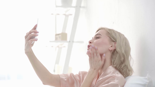 wesoły blond dziewczyna biorąc selfie na smartfonie, uśmiechając się, wysyłając pocałunki i dotykając twarzy w domu  - Materiał filmowy, wideo