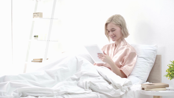 jovem loira atraente usando tablet digital e sorrindo enquanto está sentado na cama
  - Filmagem, Vídeo