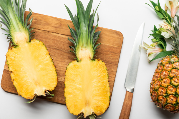 top view of cut ripe yellow pineapple on wooden chopping board near knife and Alstroemeria flowers on white background - Foto, immagini