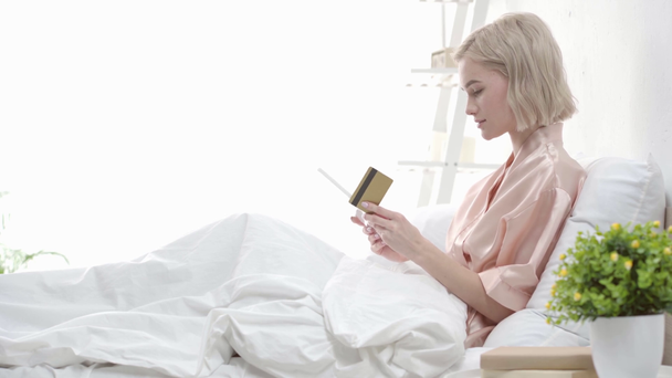 attractive blonde girl looking at credit card, using digital tablet and smiling while sitting in bed  - Felvétel, videó
