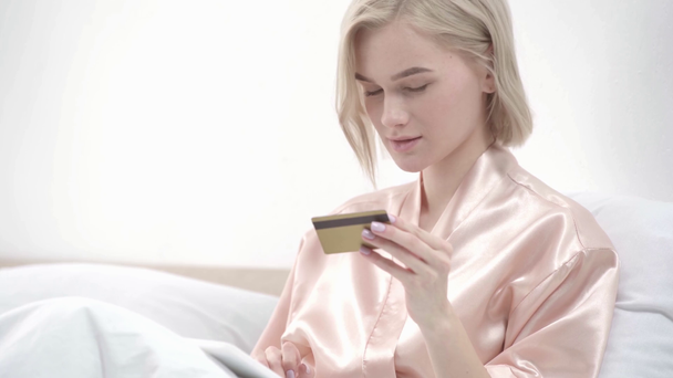 allegra ragazza bionda in possesso di tablet digitale, prendendo carta di credito e sorridente mentre lo shopping online a letto
  - Filmati, video