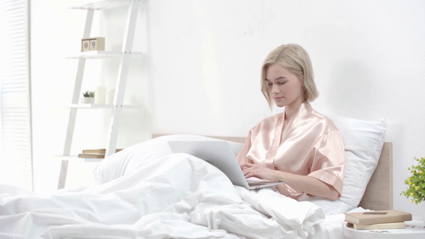 attractive blonde girl using laptop, smiling and touching face while sitting in bed  - Footage, Video