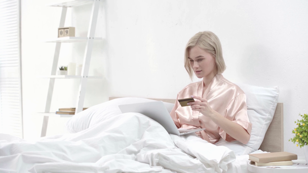 Blond dziewczyna za pomocą laptopa, biorąc karty kredytowej i uśmiechając się podczas zakupów online w sypialni - Materiał filmowy, wideo