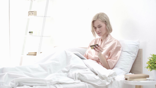Blond dziewczyna za pomocą cyfrowego tabletu, biorąc karty kredytowej i uśmiechając się podczas zakupów online w sypialni - Materiał filmowy, wideo