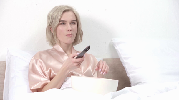 attractive blonde girl eating popcorn while holding remote controller and watching movie in bed  - Кадри, відео