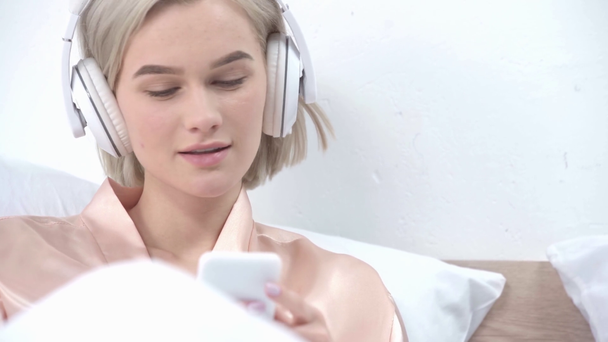 enfoque selectivo de la chica rubia feliz utilizando el teléfono inteligente y escuchar música en los auriculares en la cama
  - Metraje, vídeo