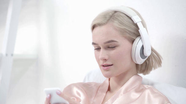heureuse fille blonde toucher écouteurs tout en écoutant de la musique et en utilisant smartphone dans la chambre
 - Séquence, vidéo