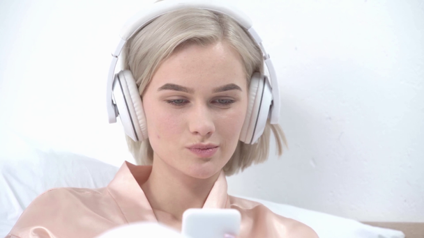весела блондинка торкається навушників, слухаючи музику і дивлячись на смартфон у спальні
 - Кадри, відео