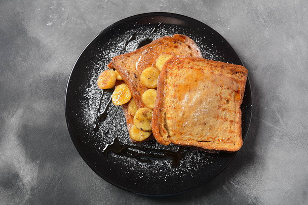 French toasts with fried plantains, dripping maple syrup, sugar powder on a black plate. French dessert concept. Sweet and delicious breakfast  - Fotó, kép