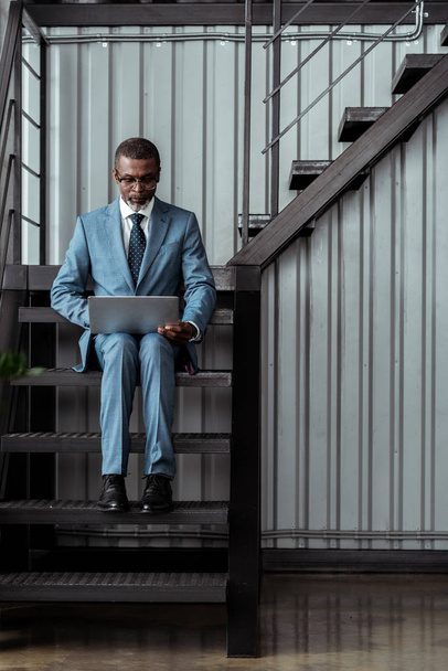 handsome african american man in glasses using laptop while sitting on stairs in office  - 写真・画像