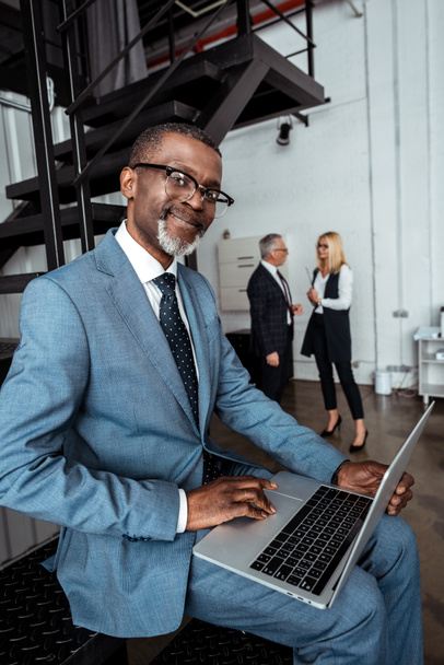 selective focus of happy african american man using laptop near partners in office  - Fotoğraf, Görsel