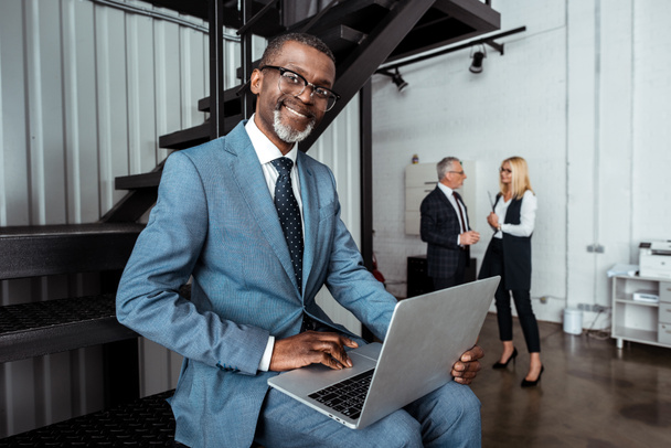 foco seletivo de empresário afro-americano alegre usando laptop perto de parceiros
  - Foto, Imagem