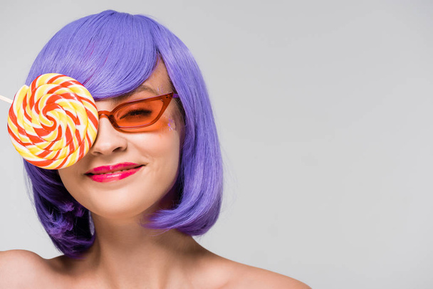 smiling girl in purple wig and sunglasses holding sweet lollipop, isolated on grey  - Foto, afbeelding