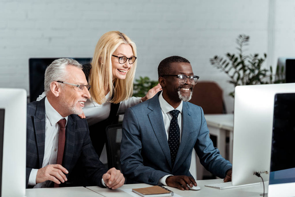 happy blonde businesswoman looking at computer monitor near multicultural businessmen  - Foto, Imagen