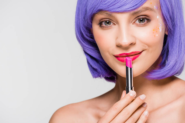 beautiful smiling girl in purple wig holding lipstick isolated on grey - Фото, изображение