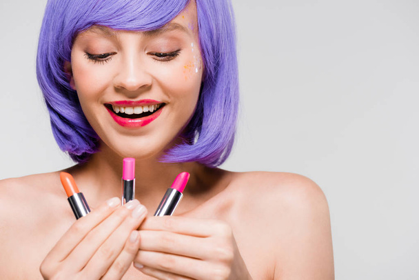 beautiful smiling woman in purple wig looking at lipsticks isolated on grey - Foto, Imagem