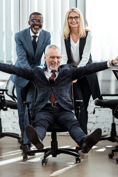 happy businessman in glasses riding on chair near multicultural coworkers  - 写真・画像