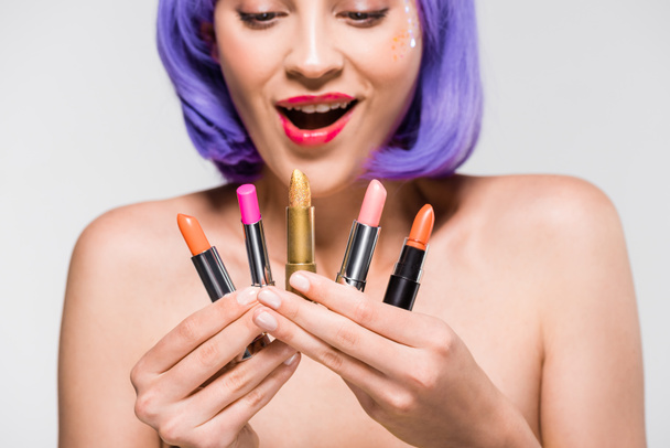 beautiful excited girl in purple wig looking at lipsticks isolated on grey - Photo, Image