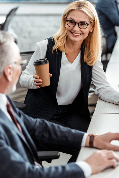 enfoque selectivo de atractiva mujer rubia sonriendo mientras sostiene la taza de papel y mirando a su compañero de trabajo en la oficina
  - Foto, imagen