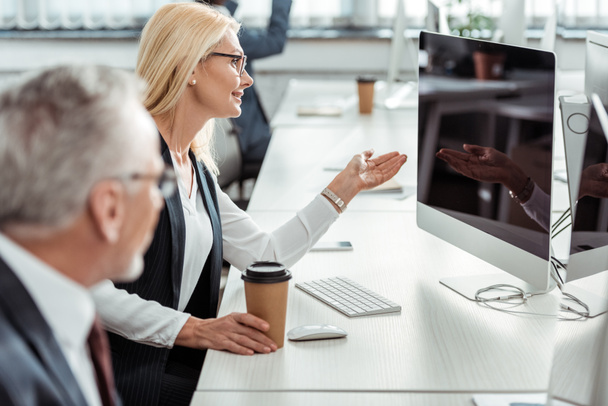 selective focus of blonde businesswoman in glasses gesturing while looking at computer monitor near coworker  - Photo, image