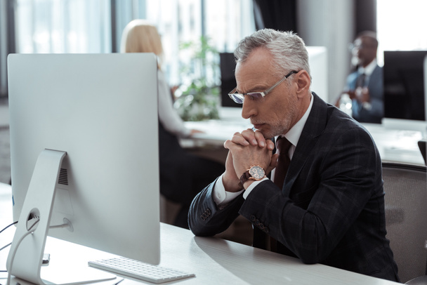 εκλεκτική εστίαση του σκεπτικού ώριμου επιχειρηματία στα γυαλιά που κάθονται κοντά στην οθόνη του υπολογιστή και τους πολυπολιτισμικούς συναδέλφους στο γραφείο  - Φωτογραφία, εικόνα