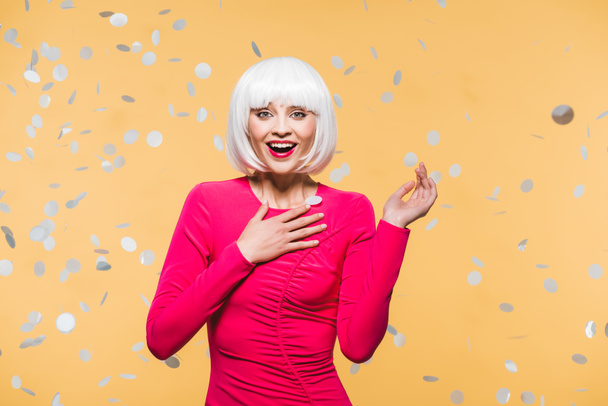 surprised girl in red dress and white wig posing with holiday confetti, isolated on yellow - Foto, imagen