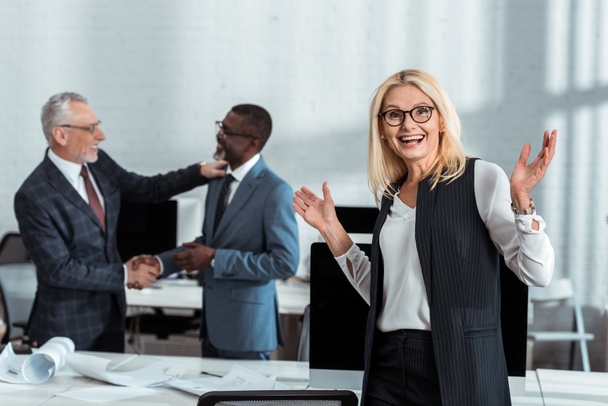 selective focus of happy businesswoman gesturing near multicultural businessmen shaking hands in office  - Фото, изображение