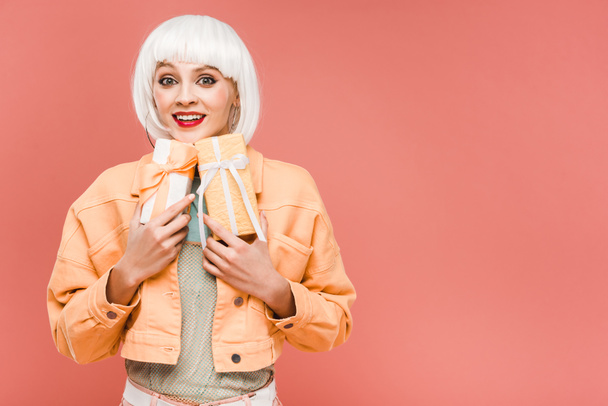 happy fashionable girl in white wig holding gift boxes, isolated on pink - 写真・画像