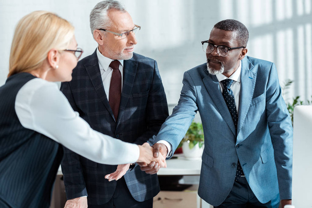 selective focus of businessman in glasses looking at handsome african american man shaking hands with blonde woman in office  - Foto, immagini