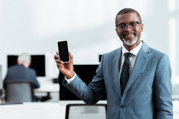 selective focus of happy african american businessman holding smartphone with blank screen in office  - Fotoğraf, Görsel