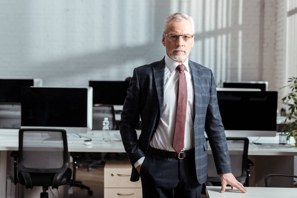 handsome businessman in glasses standing with hand in pocket in office  - Fotografie, Obrázek