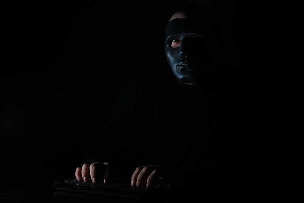 Ένας άντρας με μάσκα εκτυπώνει στο πληκτρολόγιο στο σκοτάδι. - Φωτογραφία, εικόνα