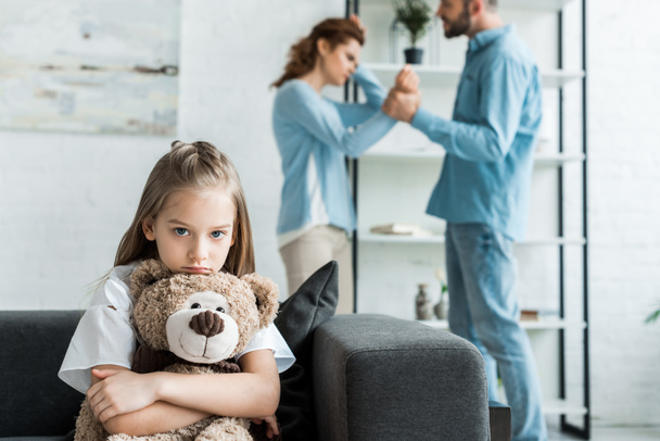 selective focus of sad kid holding teddy bear near quarreling parents at home  - Foto, afbeelding