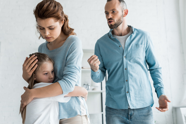 怒っている男が自宅で拳を示している間、動揺した母親は娘を抱きしめる  - 写真・画像