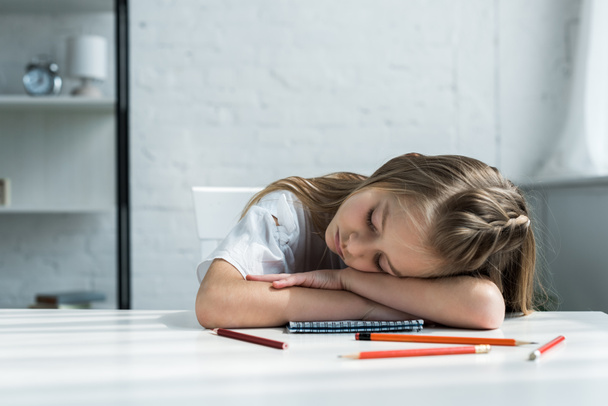 zmęczony dzieciak z zamkniętymi oczami leżąc na stole w pobliżu ołówki i notebooka  - Zdjęcie, obraz