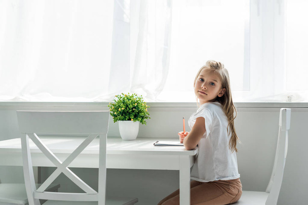 słodkie dziecko siedzi i trzyma ołówek w pobliżu notebooka w domu  - Zdjęcie, obraz