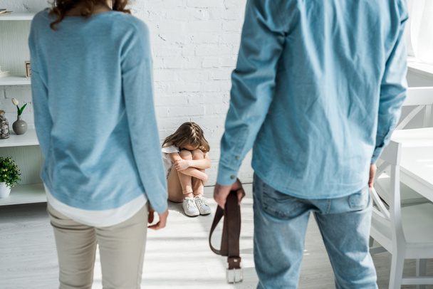 selective focus of scared kid sitting on floor near father with belt and mother  - Foto, Imagem