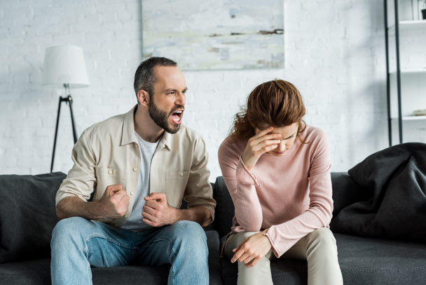 сердитый мужчина сидит на диване и кричит на расстроенную женщину
  - Фото, изображение