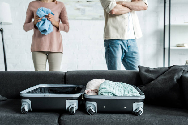 vista cortada de homem e mulher de pé perto da mala com roupas no sofá
  - Foto, Imagem