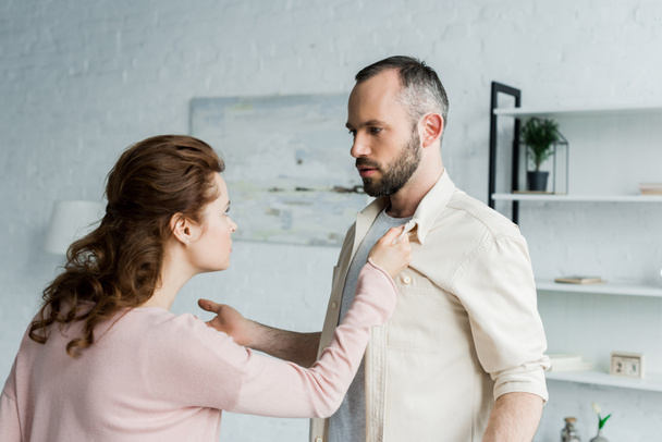 angry woman touching shirt of handsome bearded man at home  - Fotó, kép
