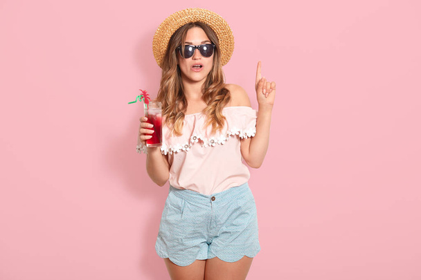 Attractive young woman in summer outfit isolated on pink background. Happy girl in sunglasses posing with straw hat and cocktail, spending hot summer days on beach, points fore finger up, has new idea - Foto, imagen