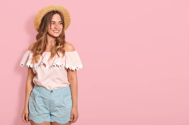 Indoor shot of pleasant looking smiling young woman stands wearing summer hat and fashionable blouse, looks dreamily aside with happy expression, isolated over pink background with free space for text - Fotoğraf, Görsel