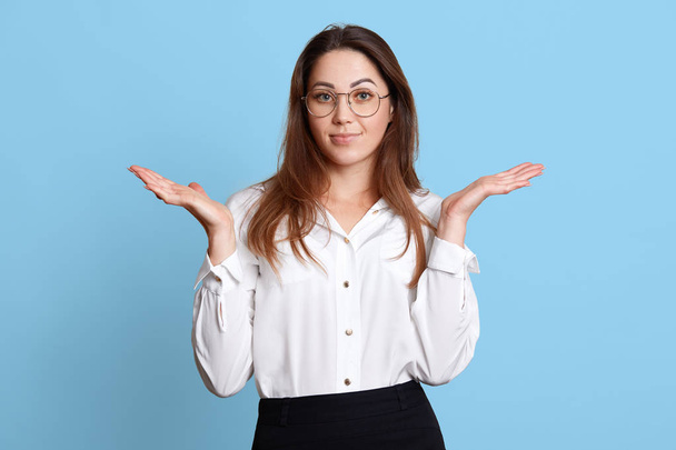 Helpless charismatic young female with long hair making gesture, raising her hands, looks confused about work, posing isolated over light blue background in studio. People and office work concept. - Photo, image