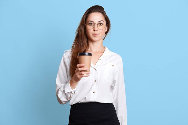 Portrait of serious business woman, wearing white shirt, black skirt and glasses, has long dark hair, holding paper cup of takeaway coffee, looks at camera, isolated on blue studio background. - Fotoğraf, Görsel